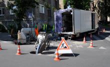 Ekipa pracowników w odzieży ochronnej przeprowadzająca prace związane z konserwacją kanalizacji na miejskiej ulicy, otoczoną pachołkami drogowymi i znakami ostrzegawczymi.
