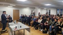 Mężczyzna w marynarce przemawia do zgromadzonych mieszkańców siedzących przy stołach, podczas spotkania w świetlicy wiejskiej.