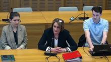 Trzy osoby siedzą przy stole konferencyjnym, a kobieta w środku mówi do mikrofonu, podczas gdy pozostałe osoby słuchają i notują.