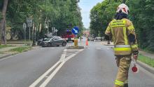 Strażak kieruje ruchem na miejscu wypadku drogowego, gdzie samochód osobowy uderzył w latarnię, a na miejscu obecne są służby ratunkowe.
