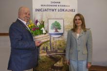 Mężczyzna trzymający kosz z wiosennymi kwiatami i figurką baranka stoi obok kobiety na tle plakatu promującego inicjatywę lokalną.