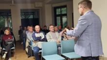 Mężczyzna w marynarce przemawia do zgromadzonych mieszkańców siedzących przy stołach, podczas spotkania w świetlicy wiejskiej.