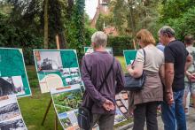 Grupa osób ogląda plansze z archiwalnymi zdjęciami i mapami podczas plenerowej wystawy historycznej. Ekspozycja ustawiona jest na sztalugach w parku, w otoczeniu zieleni. W tle widoczny ceglany budynek z czerwonym dachem.
