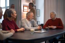 Grupa uśmiechniętych seniorów siedzi przy stole i prowadzi rozmowę w przyjaznej atmosferze. W tle widać kilku innych uczestników spotkania. Pomieszczenie jest jasne i przytulne, z dekoracjami na ścianach.