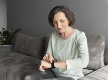 Starsza kobieta siedzi na sofie i uśmiechając się, obsługuje opaskę sportową lub smartwatch na swoim nadgarstku.