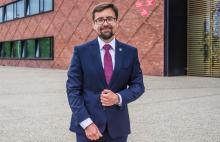 uśmiechnięty burmistrz w granatowym garniturze stoi na tle urzędu miasta i gminy Konstancin-Jeziorna