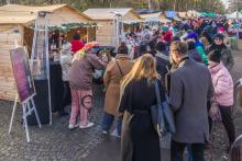 Ludzie spacerują między drewnianymi stoiskami z ozdobami i świątecznymi produktami podczas jarmarku w parku otoczonym drzewami.
