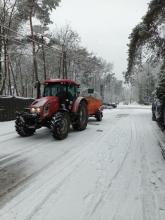 Na ośnieżonej ulicy jedzie czerwony traktor z posypywarką, wykonując zimowe prace utrzymaniowe w otoczeniu drzew i zaparkowanych samochodów.