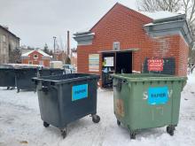 Altana śmietnikowa na osiedlu zimą, przed którą stoją przepełnione pojemniki na papier, ustawione na zaśnieżonym placu.