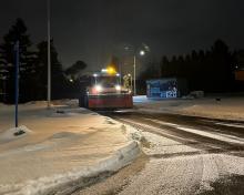 Pług śnieżny odśnieżający zaśnieżoną jezdnię, oświetloną latarniami ulicznymi, z widocznymi śladami przejazdu na mokrym asfalcie.