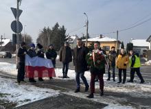 Grupa osób podczas zimowej uroczystości składa kwiaty pod pomnikiem, jedna z nich niesie wieniec, a w tle widoczna jest flaga Polski oraz zabudowa jednorodzinna przy drodze.