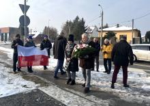 Grupa osób podczas zimowej uroczystości składa kwiaty pod pomnikiem, jedna z nich niesie wieniec, a w tle widoczna jest flaga Polski oraz zabudowa jednorodzinna przy drodze.
