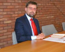 Mężczyzna w granatowym garniturze i czerwonym krawacie siedzi przy stole konferencyjnym, trzymając dłonie złożone na blacie, na tle ceglanej ściany.