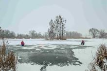 Dwóch wędkarzy siedzi na zamarzniętym stawie i łowi ryby w przeręblach, mimo cienkiej i nierównej tafli lodu pokrytej śniegiem.