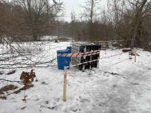 Na zaśnieżonym terenie przy zadrzewionej drodze stoją zabezpieczone taśmą ostrzegawczą pojemniki – mauzer i dwie beczki – odgrodzone drewnianymi palikami.