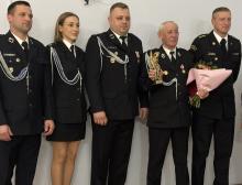 Grupa strażaków w galowych mundurach stoi obok siebie podczas uroczystości, a jeden z nich trzyma statuetkę, inny bukiet kwiatów.