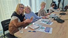 Dwie kobiety siedzą przy stole, patrzą w obietkyw i uśmiechają się. W rękach trzymają telefony komórkowe, przed nimi na stole leżą broszury, zeszyty - są na szkoleniu.