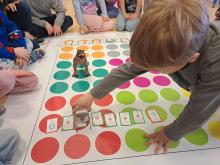 Grupa dzieci w przedszkolu siedzi na dywanie wokół dużej maty edukacyjnej z kolorowymi polami. Chłopiec na pierwszym planie ustawia małego robota oraz karty z komendami ruchu (strzałkami), planując drogę do figurki dinozaura.