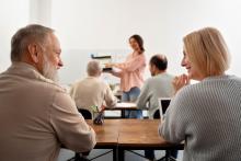 Widok zza pleców pary seniorów siedzących przy biurku w sali wykładowej. Mężczyzna z brodą i kobieta o jasnych włosach uśmiechają się do siebie. Na pierwszym planie widać organizer z przyborami do pisania. W głębi sali, na rozmytym tle, inna uczestniczka prowadzi prezentację.