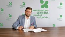 Mężczyzna w okularach podpisuje dokumenty przy stole na tle ścianki z logo „Konstancin-Jeziorna – więcej niż uzdrowisko”.