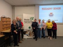 Grópa seniorów stoi w sali przy roll-upie z napisem Rada Seniorów, w tle na ścianie slajd prezentacji Rozsądny Senior