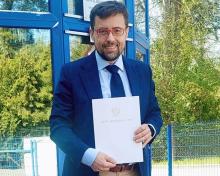 Mężczyzna w garniturze stoi przed budynkiem i trzyma w rękach dokument zatytułowany „Akt notarialny”.