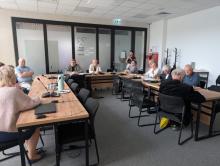 Fotografia przedstawia posiedzenie Rady Seniorów Gminy Konstancin-Jeziorna w nowoczesnej sali konferencyjnej. Grupa kilkunastu osób siedzi przy stołach ułożonych w kształt litery U. Na pierwszym planie po lewej widać tył sylwetki kobiety w różowym swetrze, a przed nią laptop i mikrofon konferencyjny na drewnianym blacie. Pozostali uczestnicy obrad, w większości seniorzy, siedzą przodem lub bokiem do obiektywu, słuchając wystąpienia. W głębi sali, za szklaną ścianą z drzwiami, na której widnieje logo gminy (
