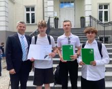 Na schodach przed budynkiem stoją cztery osoby: zastępca burmistrza gminy oraz trzej uczniowie trzymający dyplom i zielone teczki z nagrodami, laureaci konkursu wiedzy o historii Konstancina-Jeziorny.