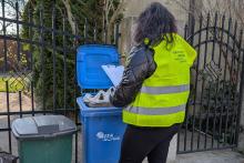 Pracownica gminy w kamizelce odblaskowej kontroluje pojemnik na odpady wystawiony przed posesją.