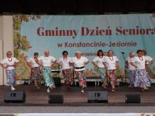 Grupa seniorek w jednakowych białych bluzkach, czerwonych koralach i kolorowych, wzorzystych spódnicach tańczy na scenie amfiteatru. W tle widać duży baner z napisem „Gminny Dzień Seniora w Konstancinie-Jeziornie”.