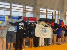 Grupa młodzieży stoi tyłem w szpalerze, obejmując się ramionami w hali sportowej. Przed nimi powieszona jest biała flaga z logo organizacji oraz napisem „CISV poland building global friendship”. W tle na szkolnych drabinkach wiszą flagi państwowe, m.in. Szwecji, Norwegii, Niemiec i Francji.