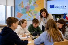 Na zdjęciu nauczycielka prowadzi zajęcia z grupą uczniów siedzących przy wspólnym stole. W tle widoczne są mapa Europy oraz ekran z logo programu Erasmus+, a także flagi Unii Europejskiej i Polski. Uczniowie wspólnie pracują nad zadaniem w przyjaznej atmosferze.