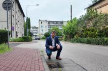 Burmistrz kuca przy wpuście kanalizacji deszczowej na osiedlowej ulicy, wskazując element infrastruktury przeznaczonej do modernizacji, a w tle widoczne są budynki mieszkalne i zieleń.