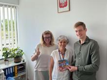 Na zdjęciu trzy osoby stoją w urzędzie i prezentują pocztówkę z Walencji zgłoszoną do konkursu „Pocztówka z wakacji”. Jedna z osób trzyma kciuk uniesiony w geście aprobaty, a dwie pozostałe wspólnie prezentują widokówkę do obiektywu.