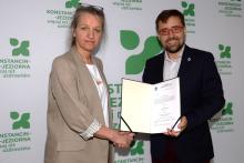 Na zdjęciu kobieta (dyrektor Ewa Winek) i mężczyzna (burmistrz Michał Wiśniewski) stoją przed ścianką z logo gminy Konstancin-Jeziorna. Uściskiem dłoni potwierdzają wręczenie aktu powierzenia stanowiska dyrektora. Mężczyzna trzyma otwartą teczkę z widocznym dokumentem i uśmiecha się do obiektywu.