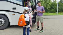 Dzieci z plecakami wsiadają do autokaru przed wyjazdem na wakacyjny wypoczynek w Zakopanem. Opiekun sprawdza listę uczestników.