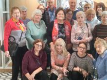 Grupa kilkunastu seniorek i kilku seniorów pozuje do wspólnego zdjęcia we wnętrzu budynku. Część osób stoi, a cztery kobiety siedzą lub klęczą z przodu.