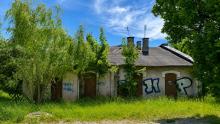 Zaniedbany budynek dawnej parowozowni z zamurowanymi oknami i graffiti na ścianach. Obiekt otaczają wysoka trawa i drzewa