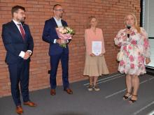 Cztery osoby stoją przed ceglaną ścianą podczas uroczystego podziękowania. Jedna z kobiet trzyma dyplom, mężczyzna bukiet kwiatów, a druga kobieta przemawia do mikrofonu