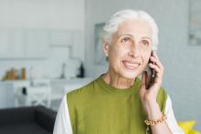 Uśmiechnięta seniorka o siwych włosach rozmawia przez telefon komórkowy. Kobieta przebywa w jasnym, domowym wnętrzu