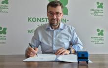 Burmistrz siedzi przy biurku i podpisuje dokumenty, a na stole obok znajdują się pieczątka oraz dokumentacja.