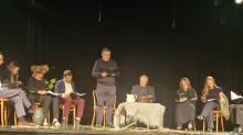 Zdjęcie z czytania Dziadów na ciemnej scenie. Grupa osób siedzi na krzesłach wzdłuż sceny, trzymając w rękach egzemplarze tekstu, podczas gdy mężczyzna na środku czyta na stojąco. Przed nimi na podłodze leżą rozrzucone gałęzie, a pośrodku stoi mały stolik przykryty jasnym obrusem z dzbankiem i misą.