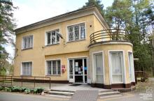 Budynek przychodni wśród drzew, z żółtą elewacją, dużymi oknami z kratami oraz zaokrąglonym wejściem z podjazdem dla osób z niepełnosprawnościami.