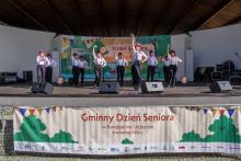 Występ artystyczny seniorów na scenie amfiteatru podczas Gminnego Dnia Seniora w Konstancinie-Jeziornie. Grupa tancerzy w białych koszulach, ciemnych spodniach, czerwonych szelkach i kapeluszach wykonuje układ taneczny z uniesionymi rękami. Z przodu sceny wisi duży baner z napisem „Gminny Dzień Seniora w Konstancinie-Jeziornie”.
