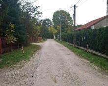 Droga gruntowa z kruszywa, biegnąca między ogrodzonymi posesjami, z drzewami i zielenią po obu stronach oraz widoczną infrastrukturą energetyczną.