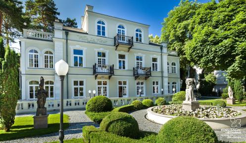 Villa la Fleur Trzykontygnacyjna willa – jasna elewacja, dużo okien, płaski dach. Przed budynkiem stoi fontanna. Dokoła rosną wysokie drzewa.