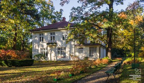 Widok na willę Świt. Biały budynek na planie kwadratu z czerwonym dachem, duża ilością okien. Wokół willi rosną wysokie drzewa. Zdjęcie zrobione w jesienny dzień.