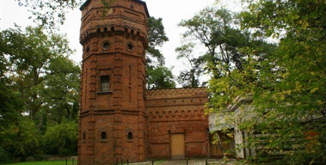 Zabytkowa budowla z czerwonej cegły z wysoką wieżą po prawej stronie.  Kilka małych okien.  Dokoła wysokie drzewa.