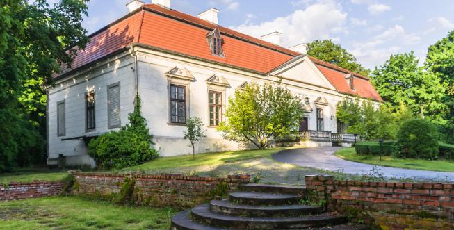 Prostokątny budynek (dwór) z jasną elewacją, dużą ilością okien, z pomarańczowym dwuspadowym dachem. Dokoła rosną wysokie drzewa. Do dworu z ogrodu prowadzą szerokie, betonowe schody.