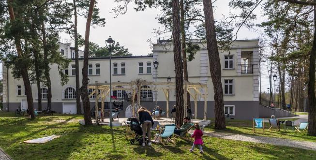 W głównej części zdjęcia znajduje się rodzinka (rodzice i dwójka dzieci), którzy siedzą na leżakach w wiosenny dzień. Za nimi widoczna jest drewniana konstrukcja pergoli, oraz duży budynek, w którym mieści się siedziba Konstancińskiego Domu Kultury. 